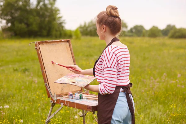 Visão traseira da jovem artista em pé na frente do caderno de esboços com escova e paleta de cores, pinturas imagem na natureza no verão, vestindo roupas casuais, tem cabelo claro. Conceito de arte e criação . — Fotografia de Stock