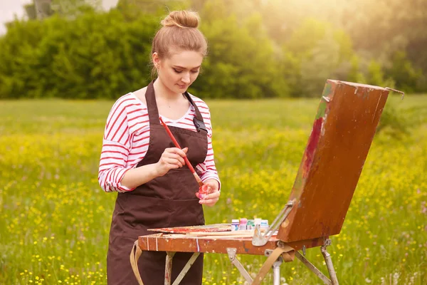 Retrato de bela fêmea olhando para amostras de aquarela em convas, cores misturadas, tem expressão facial concentrada, mulher usa roupas casuais, tem cabelo claro e maço. Conceito de arte e criação . — Fotografia de Stock