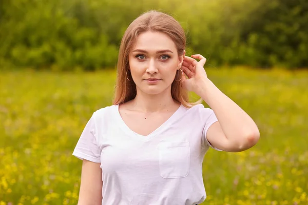 Внешний снимок привлекательной молодой красивой блондинки волосы стройная девушка, в повседневной футболке позируя на открытом воздухе, глядя прямо на камеру, имеет спокойный взгляд, изолированы на зеленом фоне луга травы . — стоковое фото