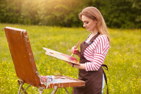 Imagem de uma jovem mulher desenhando quadro sobre tela, usando sketchbook para desenhar na natureza, perfil menina pintor com escova e paleta de tintas de trabalho inspirado na paisagem do prado. Conceito de estilo de vida — Fotografia de Stock