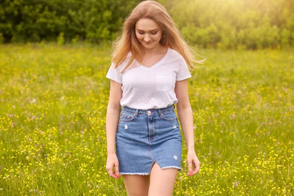 Nuestra foto puerta de la joven hermosa chica caucásica en camiseta blanca y falda de mezclilla azul posando contra el atardecer en el prado, mirando hacia abajo, atractiva hembra mirando hacia abajo. Concepto de estilo de vida y felicidad . — Foto de Stock