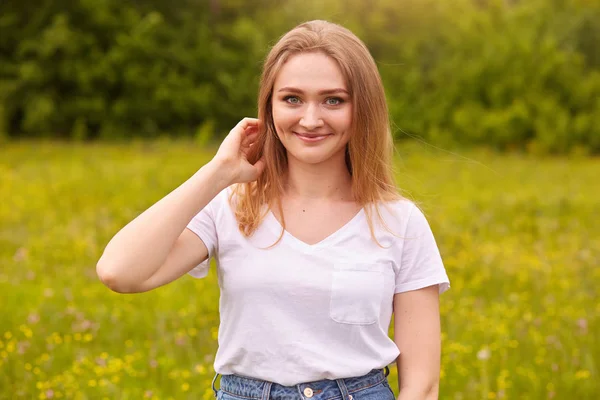 Lady with fair hair and blue eyes, looks romantic and fresh, posing in meadow, touching her hair, wearing casual white t shirt, spending summer days in nature with her boyfriend. Lifestyle concept.