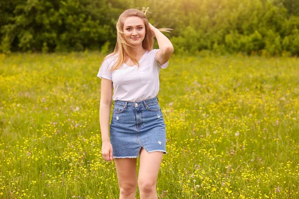 Photo extérieure de jolie femme blonde portant un t-shirt blanc décontracté et une jupe en jean, posant dans la prairie, touchant ses cheveux blonds, regardant la caméra avec une exprassion faciale agréable. Concept de bonheur . — Photo