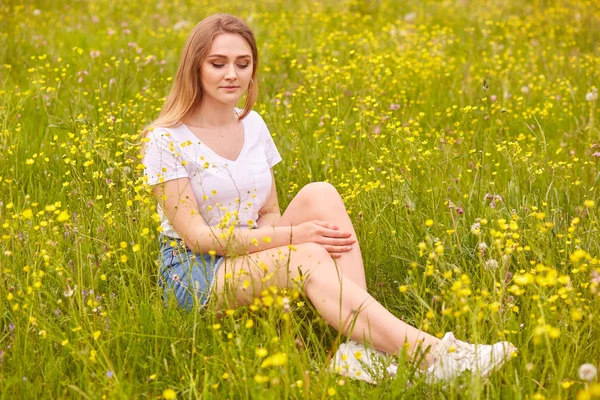 Close up retrato de jovem mulher loira modelo sentado na grama, posando em branco casul t-shirt e saia jeans, tem expressão romântica, situando-se no chão cercado flores de campo e olhando para baixo . — Fotografia de Stock