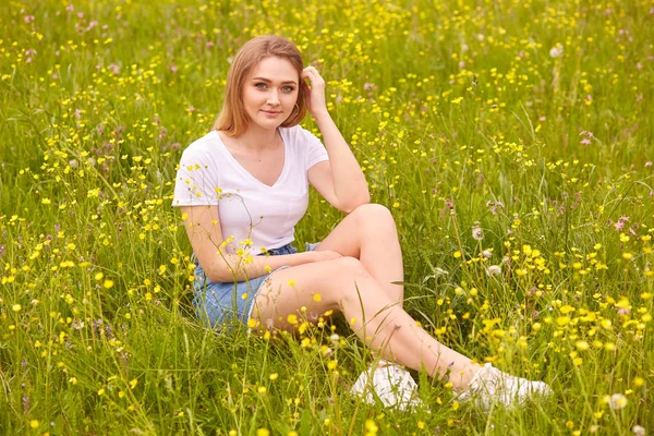 Imagem da bela menina sorridente muito feliz sentado no prado de verão verde e olhando diretamente para a câmera, atraente fêmea vestindo t-shirt branca casual e saia de ganga, senhora toca suavemente seu cabelo — Fotografia de Stock