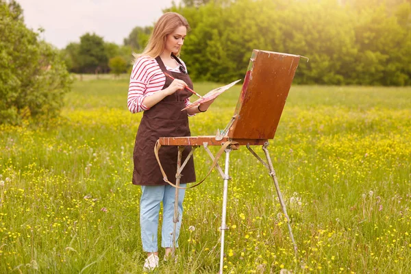 Außenaufnahme eines kreischenden Mädchens mit Pinsel und Farbpalette, das auf einer Wiese in schöner Landschaft arbeitet, Frau mit gestreiftem Hemd, lässiger Hose und brauner Schürze. Kunst und Kreationskonzept. — Stockfoto