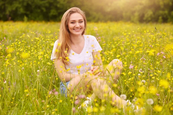 Yaz otlağında poz veren genç moda kadını. Doğanın en gözde kızı. Sarı tarla çiçeklerinin arasında oturan mutlu gülümseyen kadın, mutluluk ve mutluluğu ifade eder. Yaşam tarzı kavramı. — Stok fotoğraf