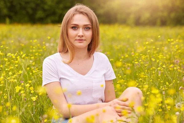 Szőke lány ült körül sárga mező virágok, már hosszú haja, szőke lány pózol a szabadban a réten treesin hátsó helyzetben, felöltözve alkalmi fehér póló, kiadások nyári napon a természetben. — Stock Fotó