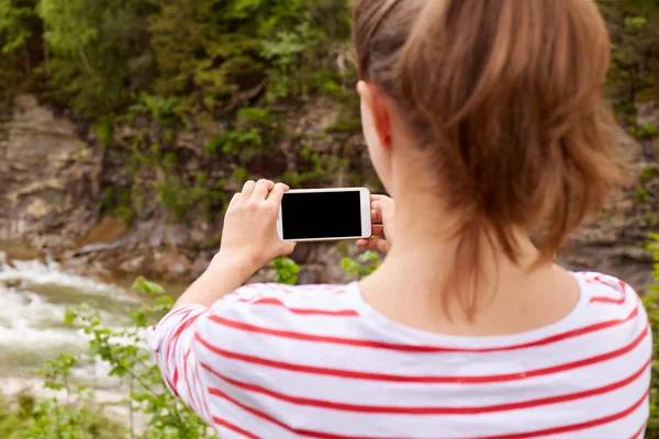 Fille voyageur prend des photos de rivière luxuriante de montagne dans le ravin sur smartphone, mise au point sélective, enjolivant paysages natures, femme portant chemise dépouillée, avec queue de cheval. Personnes et concept de voyage . — Photo