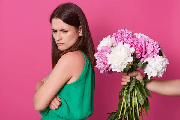 Kaşlarını çatmış yüzü çiçeklerden yüz çeviren çekici esmer genç kadın, katlanmış kolları ile stüdyoda pembe arka plan üzerinde izole ayakta, kenara bakarak, üzgün yüz ifadesi olan.