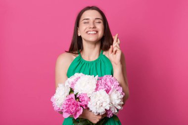 Mutlu sevimli kadın düz kapalı gözler ve barışçıl yüz ifadesi ile ayakta, parmaklarını geçiş, hayal, daha iyi umuduyla, beyaz ve pembe çiçekler bir el ile buket tutarak.
