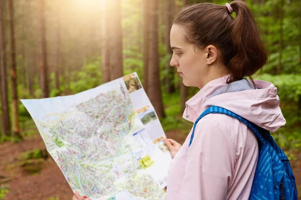 年轻的热情的旅行者站在森林中间,围绕绿色和树木,拿着地图,仔细看它,选择更好的旅行路线,寻找出路从森林. — 图库照片