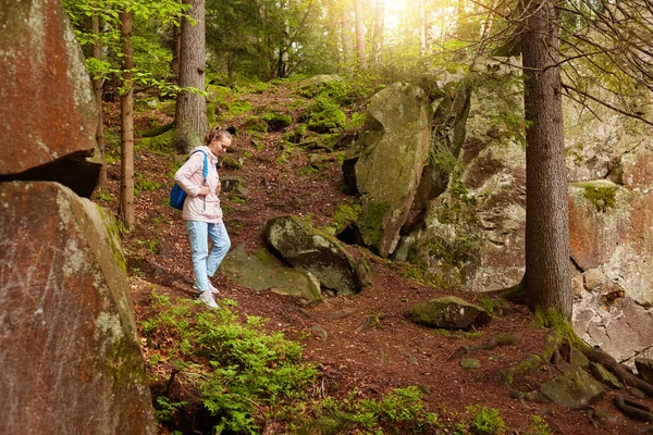 Schlanke, ernsthafte Frau, die den Berg hinuntergeht, sich von der Natur inspirieren lässt, durch steinige Hänge, Waldbäume wandert, Urlaub macht, bekannten Pfaden folgt, lässig gekleidet ist. Reisekonzept. — Stockfoto