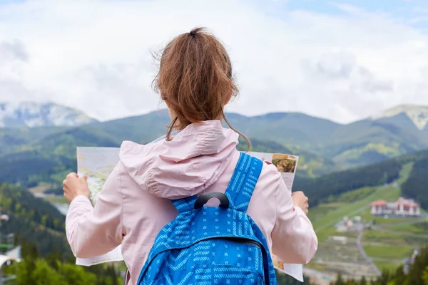 Zadní pohled na cestování stojící na vrcholu kopce s mapou, která se začíná pohybovat, a dívá se na nádhernou horskou krajinu, nosí sako, modrý batoh, s koňským ocasem. Koncepce cestování. — Stock fotografie