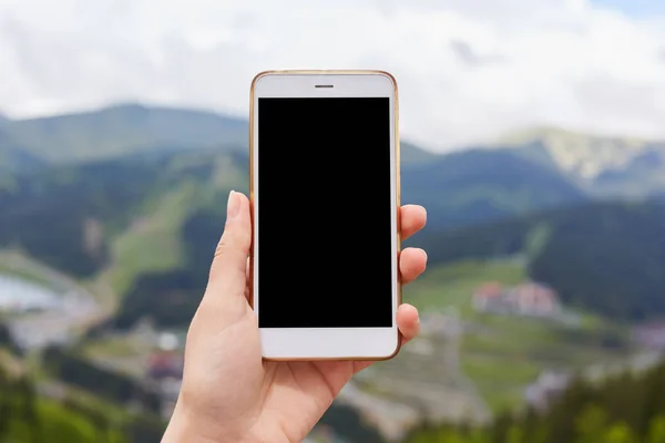 Image extérieure d'une main tenant et montrant un smartphone blanc avec écran noir blanc de bureau avec des montagnes vert flou en arrière-plan, téléphone mobile éteint. Copyspace pour la publicité . — Photo