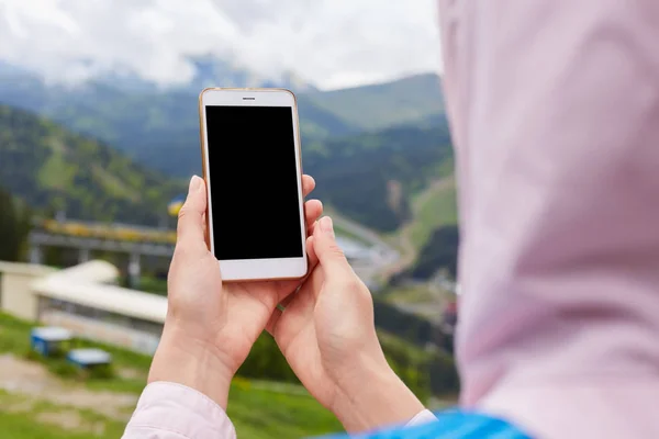 Image of faceless woman holding white smart phone with blank desktop screen outdoor, posing over green mountains background, female wearing rosy jacket. Traveling and active recreation concept. — ストック写真