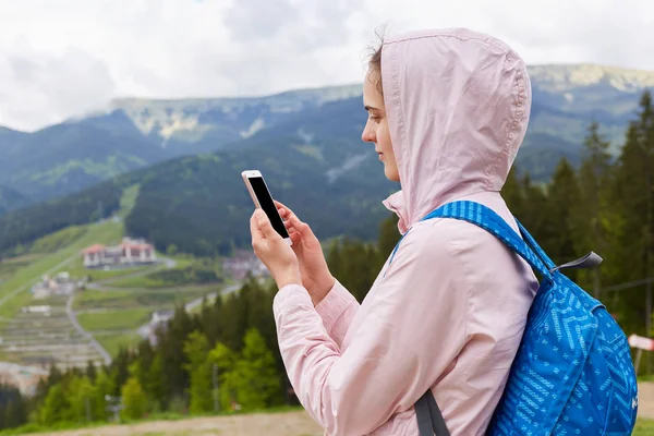 Portrait de profil rapproché de jeune femme randonneuse avec sac à dos bleu prenant des photos de beaux paysages de montagne au printemps, veste rose avec capuche. Concept de voyage et de repos actif . — Photo