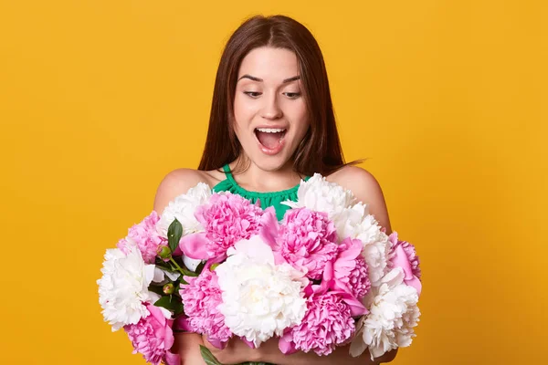 Close up portrtait of beautiful surprised romantic young woman, posing with open mouth, smiling girl holding bouquet of white and pink peony flowers. Indoor, studio shot, isolated on yellow background — Stock Photo, Image