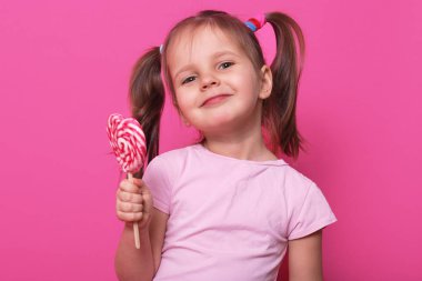 Rahat t shirt, mutlu küçük kız büyük şeker lolipop tutan gül giyen sevimli çocuğun portre yakın, zevkle yüz ifadesi büyüleyici, stüdyoda pembe arka planda izole poz.