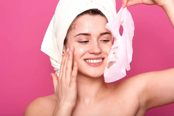 Atrakcyjna delikatna panienka stojący na białym tle na różowym tle w studiu, trzymając maskę kosmetyczną w jednej ręce, dotykając jej twarzy, uczucie jedwabistej skóry po zabiegach kosmetycznych, jest zachwycona. — Zdjęcie stockowe