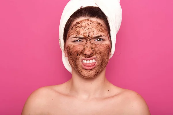 Potret perempuan yang tidak puas dengan masker coklat yang diaplikasikan di wajahnya, melihat langsung ke kamera, menunjukkan lidahnya, tidak ada kesenangan melakukan prosedur kecantikan, setelah membungkus handuk di kepala . — Stok Foto