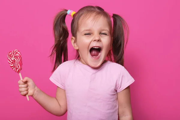 Studio shot dari sedikit menarik membuka mulut anak secara luas, melihat langsung ke kamera dengan kegembiraan, memegang lolipop terang di tangan, mengenakan santai, terisolasi atas latar belakang studio merah muda . — Stok Foto