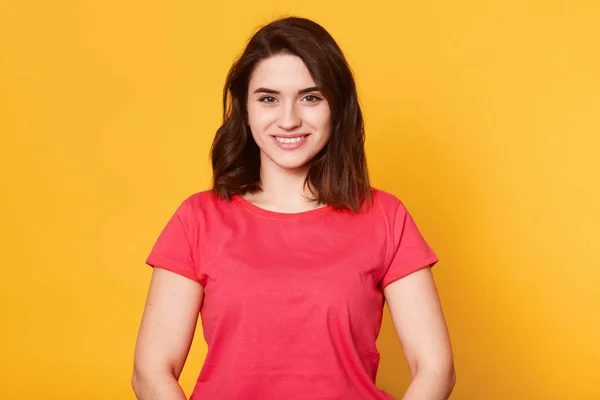 Adorable mujer agradable vistiendo casual camiseta roja posando aislado sobre fondo amarillo en el estudio, mirando directamente a la cámara, con una sonrisa sincera en su cara, luciendo agradable. Concepto de emociones . —  Fotos de Stock