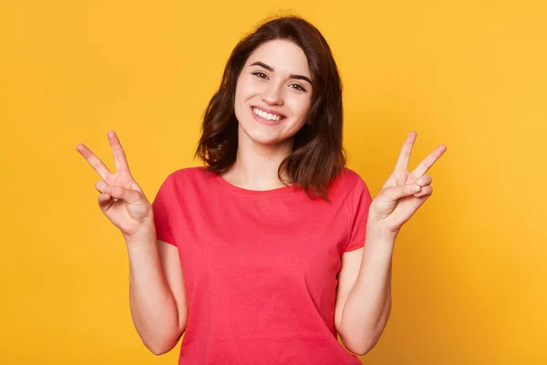 Estudio interior plano de mujer enérgica bien parecido stnading aislado sobre fondo amarillo, haciendo gestos, levantando los brazos, mostrando dos dedos, mirando encantado y pacífico. Concepto de emociones . — Foto de Stock