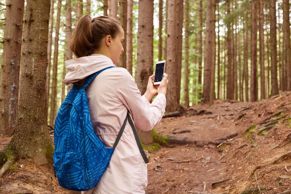 Profile of hiker woman wandering in forest, walking up slope, holding smartphone in both hands, switching it on, looking for connection, wearing pink jacket and blue backpacker. Travelling concept.