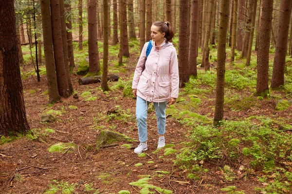 Outdoor shot van enthousiaste toerist met Camping Tour, op zoek naar het juiste pad, ziet er blij, het dragen van jeans, roze jas en blauwe backpacker, met pony staart. Vrijetijdsbesteding concept. — Stockfoto