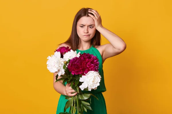 Studio shot de hermosa chica morena tiene un gran ramo de peonías borgoña en la mano, mujer reflexiva con flores mantiene la mano en la cabeza, posa sobre el fondo amarillo del estudio. Copiar espacio para promoción — Foto de Stock