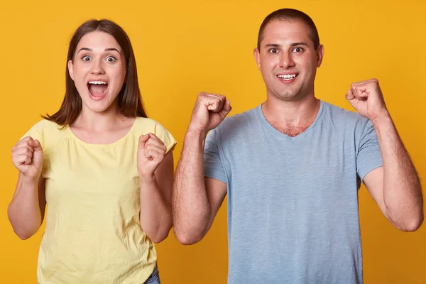 Indoor shot of happy couple marrieds clenches fists, have excited facial expressions, celebrate success, dressed in casual t shirts, isolated over yellow studio background. Relationship concept. — Stock Photo, Image