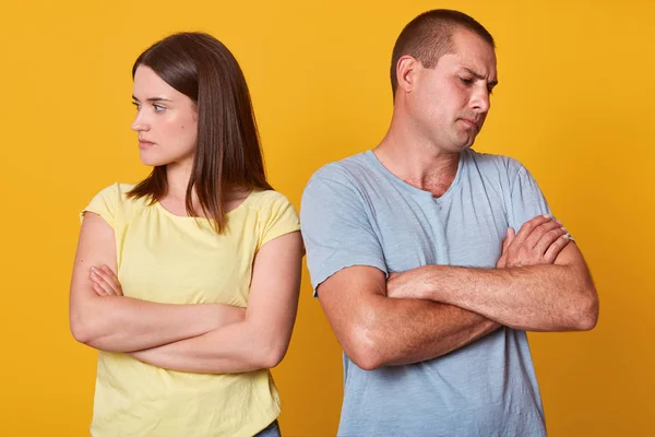 Photo of unhappy couple, man and woman, standing with face turned away from each other, with arms folded, marrieds being in quarrel, has conflict, posing isolated over yellow studio background.