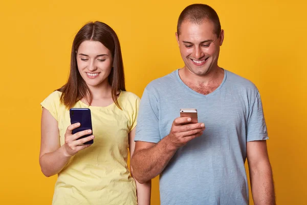 Photo of positive excited man and woman, looking at their smartphones screens with happy expressions, both using mobile phones isolated over yellow background, checkig their social networks, — Stock Photo, Image