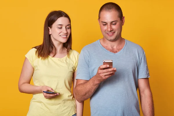Handsome man with mobile phone in hand, checking his social network, stands near cute playful brunette woman which looking in his smart phone. Positive couple isolated over yellow background. — Stock Photo, Image