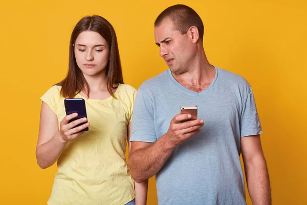 Indoor shot of young marrieds with mobile phones, demonstrates posted photos in social networks, browses in internet. Calm attentive brunette woman showing something at her screen to husband. — Stock Photo, Image