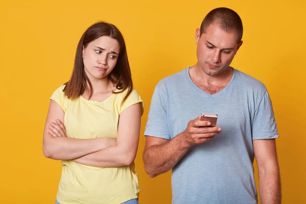 Disappointed young girl tired of being alone, looking at her boyfriend focused on his device, standing with folded arms. Busy handsome man paying no attention to his girlfriend, net addicted. — Stock Photo, Image