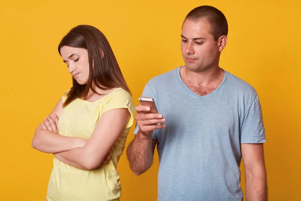 Indoor studio shot of woman being resentful to his boyfriend behavior, looking aside with folded arms, man interested in social medias, having net addiction, isolated over yellow background. — Stock Photo, Image