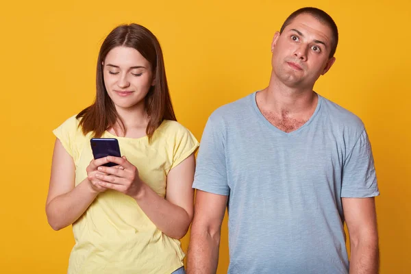 Studio shot of young couple, attractive female texting message on smartphone, male with thoughtful facial expression looking up, woman and man wearing casually, posing isolated over yellow studio wall — Stock Photo, Image