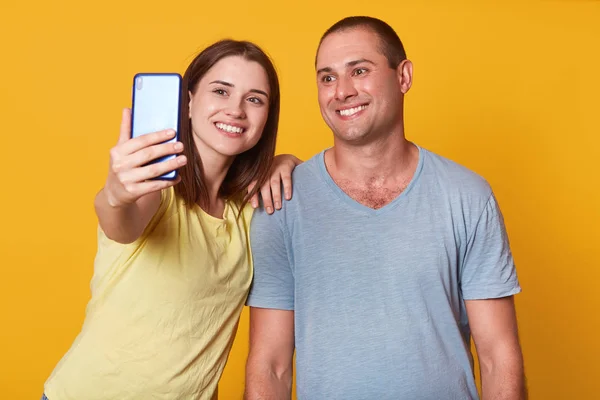 Indoor shot of beautiful young couple looking at camera of cell phone, make selfie portrait, wearing gray and yellow t shirts, posing indoor, having fun together, isolated over studio background.