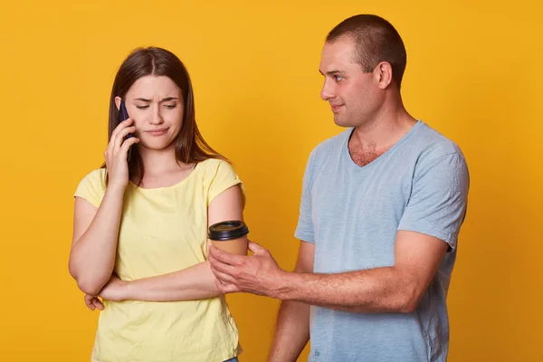 Sad woman looks down with upset expression while speaking via smart phone with her friend, standing with her husband against yellow studio wall, male offers to drink coffee. Relationship concept. — Stock Photo, Image