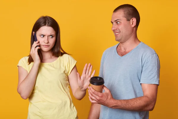 Indoor shot of sad woman looks with upset expression at guy offers her takeaway coffe, has upset facial expression, talks via cell phone, standing against yellow studio wall, marrieds wear casually. — Stock Photo, Image