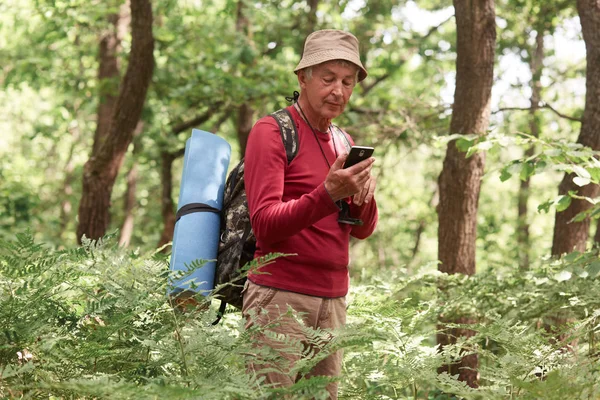 有经验的旅行者一手拿着智能手机,使用设备定位,独自,有指南针和睡眠垫,戴着帽子,裤子,运动衫,背包。旅行理念. — 图库照片