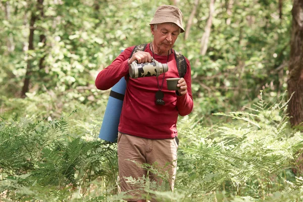 Energetic senior man having lunchtime in forest during camping trip, holding thermos, pouring drink, spending time around nature, enjoying atmosphere, having all necessary equipment to stay in forest. — 图库照片