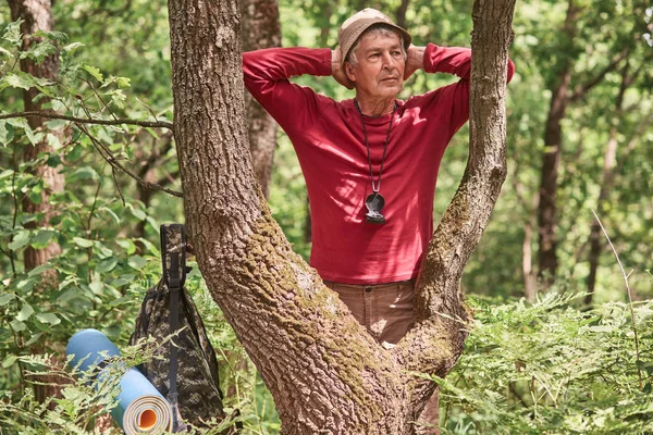 Ritratto di vecchio turista attivo che fa un giro nella foresta, si riposa, tocca la testa con entrambe le mani, guarda da parte, sta in piedi vicino all'albero, ha tutti i beni nelle vicinanze. Concetto di vita attivo . — Foto Stock