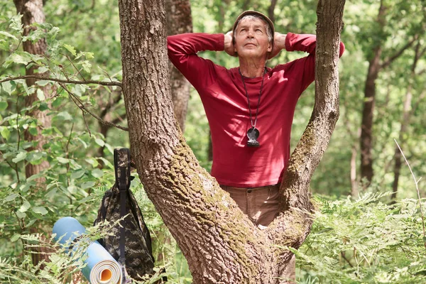 Escursionista uomo escursioni nella foresta, si ferma per riposare dopo lunghe ore zaino in spalla, sano anziano persona sportiva con lo zaino, si trova vicino all'albero e gode della natura selvaggia, indossa con disinvoltura. Concetto riposo attivo . — Foto Stock