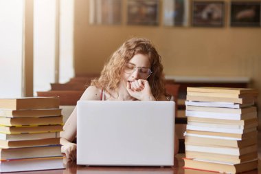 Kütüphanede masada oturan yorgun bayan, esneme sırasında yumruk ile ağzını kaplayan, durmadan çalışan, önemli bir projeye hazırlanıyor, yapılacak çok fazla görev, etrafında birçok kitap olması.