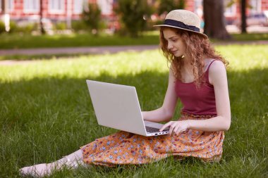 Dizüstü bilgisayarını tutan, uygun bilgileri arayan, özenli olan, yeşil alanda çimenlerin üzerinde oturan, temiz havadan ilham alan özenli meşgul öğrencinin görüntüsü. Öğrenci yaşam kavramı.