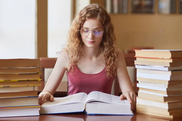 Portrait of hard working thoughtful college student with fair curly hair reading huge book, doing research, preparing for her lessons, looking attentively, having many tasks to do. Student life. — Stock Photo, Image