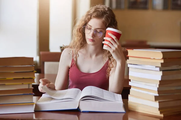 Cansada estudiante universitaria determinada en busca de información apropiada para su proyecto, leyendo textos, poniéndose papercup de bebida en la cabeza, vistiendo camisa roja y anteojos, trabajando sin parar . — Foto de Stock
